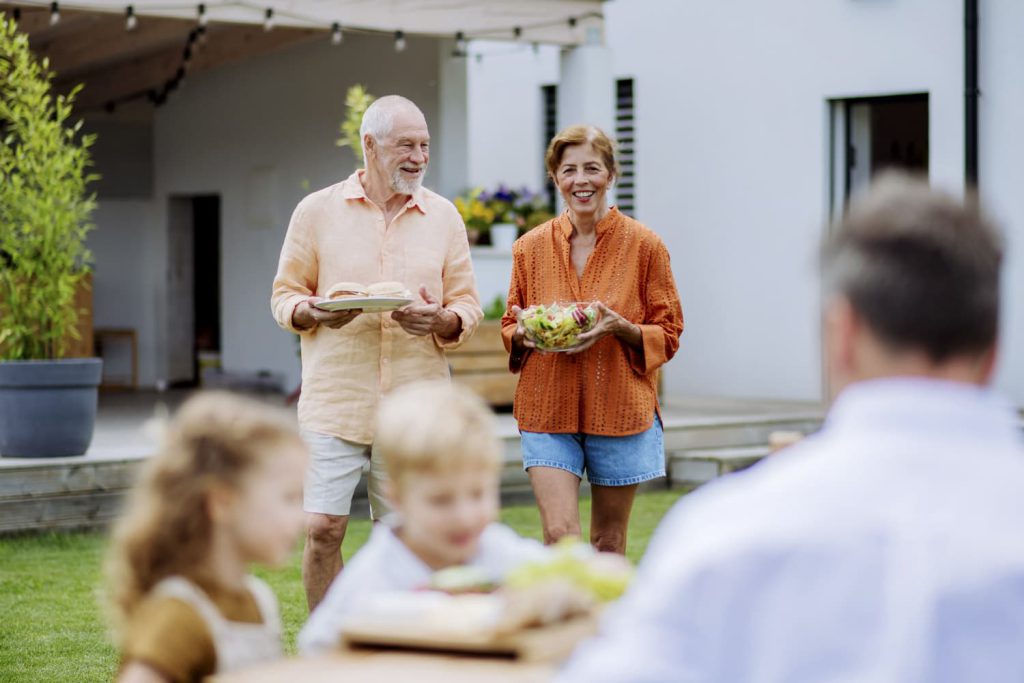 An older couple carrying plates of food while smiling at a family gathering outdoors with children seated at a table in apache junction az