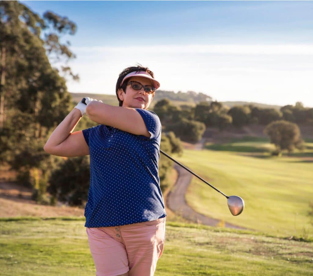 A woman in a visor and sunglasses swings a golf club on a green golf course with rolling hills in the background in apache junction az