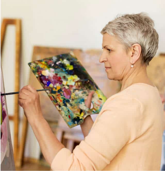 A woman with short gray hair holding a colorful palette and brush while painting on a canvas indoors in apache junction az