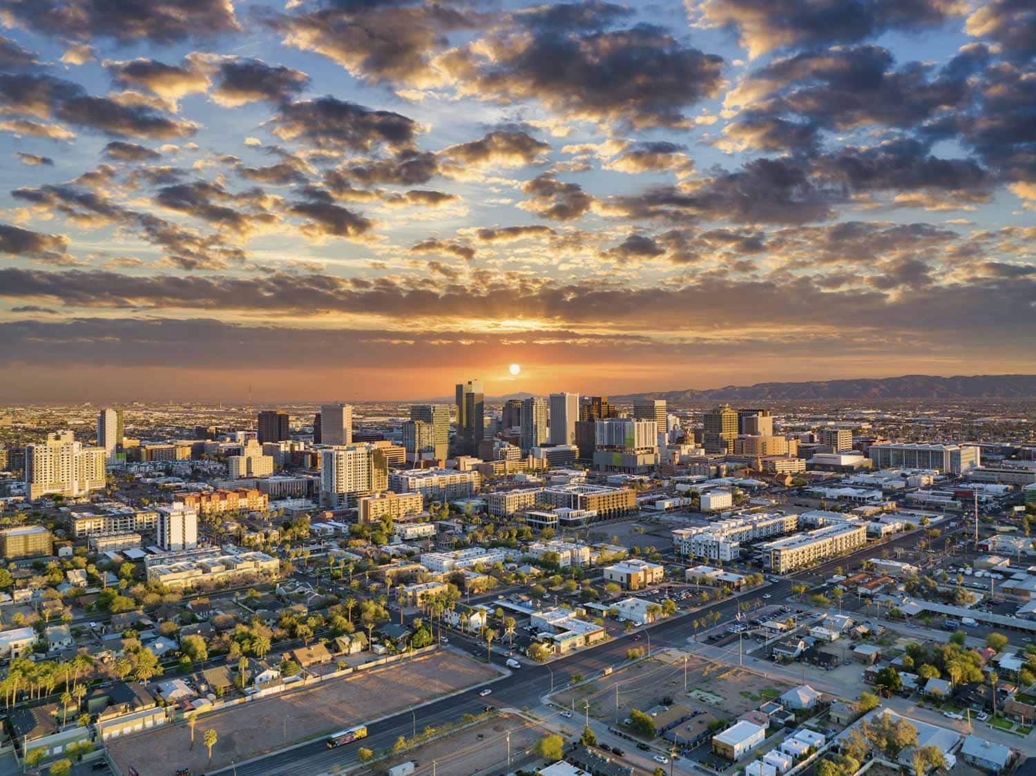 An aerial view of a city skyline at sunset with tall buildings and scattered clouds lit by the setting sun, creating a vibrant orange and blue sky in apache junction az