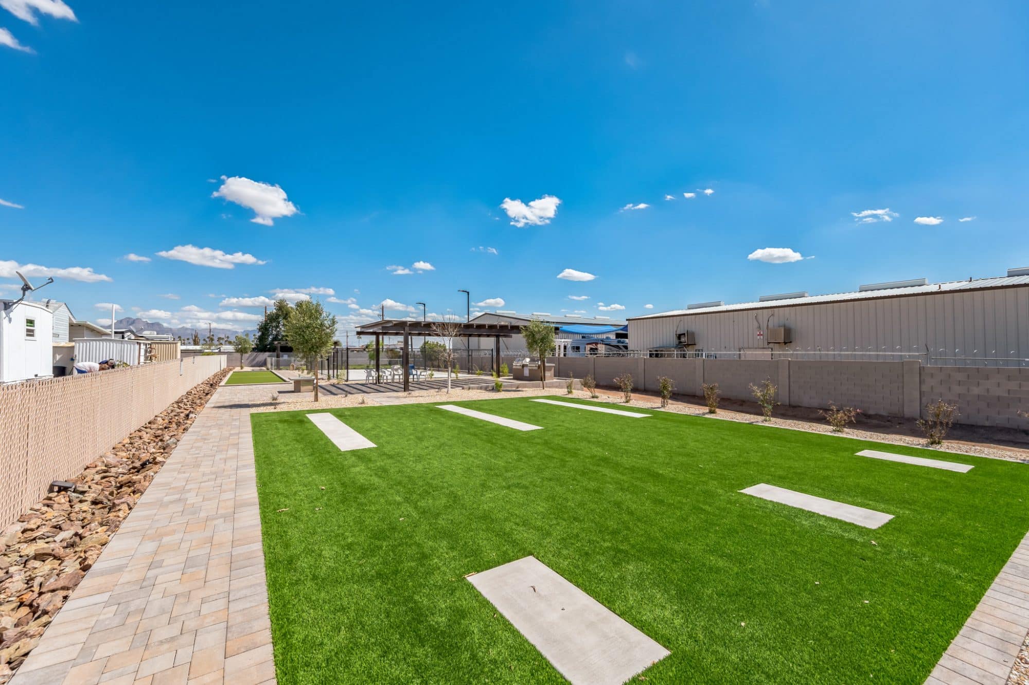 A well-maintained grassy lawn with concrete pads, surrounded by a paved walkway and a shaded pergola under a bright blue sky in apache junction az